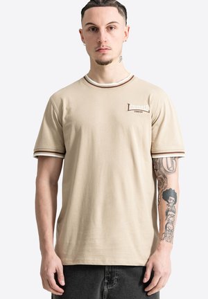 Młody mężczyzna stoi przodem, ubrany w beżowy t-shirt Lonsdale London z brązowymi paskowanymi mankietami i graficznymi tatuażami na prawym przedramieniu.