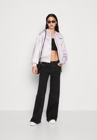 Giacca bomber in satin lavanda, top corto nero, jeans flair neri, sneakers bianche e piccola borsa nera. La modella indossa occhiali da sole.