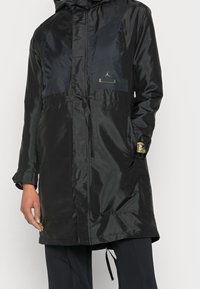 Parka imperméable noire avec capuche, finition brillante, panneaux diagonaux bleu marine foncé, taille avec cordon élastique et détails de logo dorés.