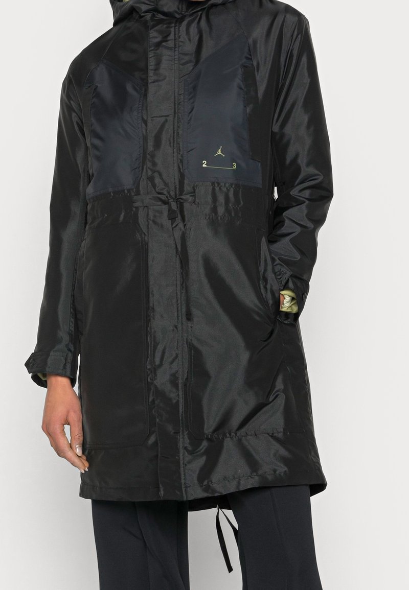 Parka imperméable noire avec capuche, finition brillante, panneaux diagonaux bleu marine foncé, taille avec cordon élastique et détails de logo dorés.