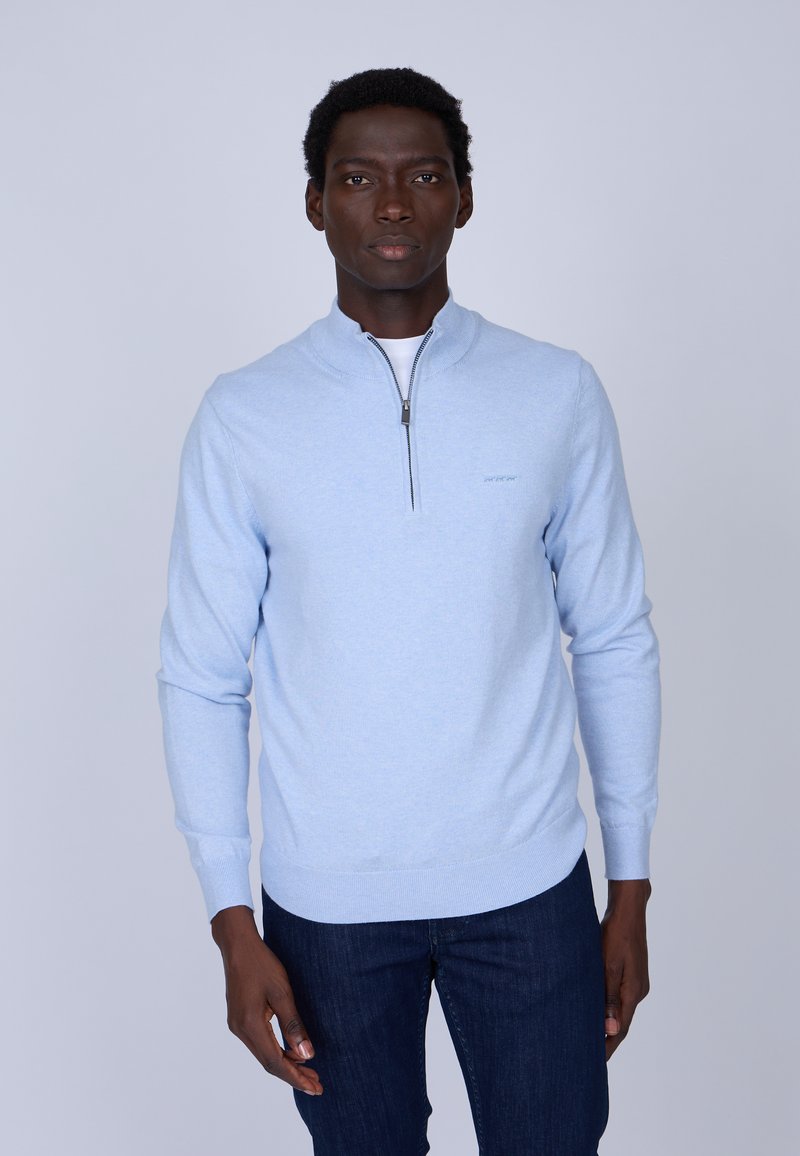 Sweater zippé bleu clair en tricot doux avec col, présentant une fermeture éclair argentée et des poignets ajustés. Porté sur un t-shirt blanc avec un jeans foncé.