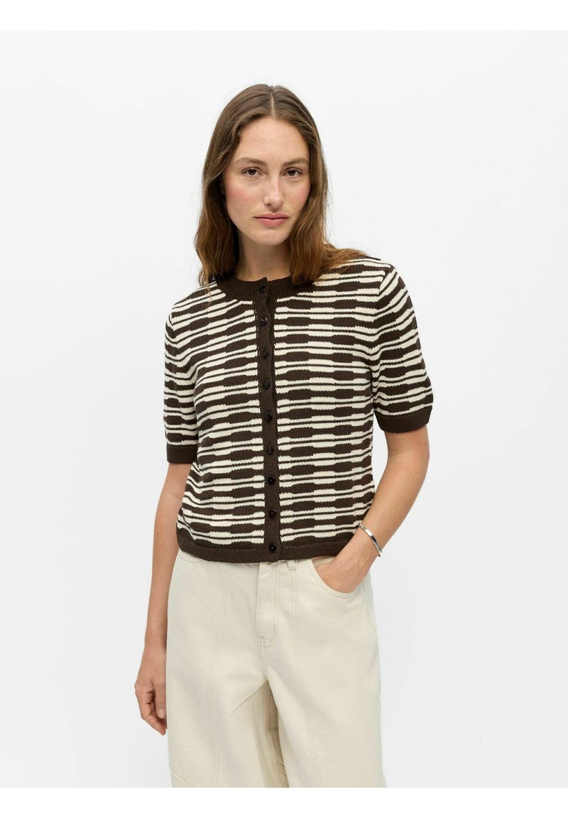 Femme portant un haut à manches courtes boutonné avec un motif horizontal marron et blanc et un pantalon de couleur claire sur un fond uni.