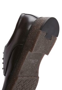 Chaussure en cuir noir avec une semelle en caoutchouc texturée. La semelle présente un motif de bande de roulement distinct et un bord arrondi. Décorée de clous sur le côté.