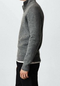 Grau gestrickter Pullover mit hohem Kragen, gerippter Textur und langen Ärmeln. Eng an den Körper geschnitten und über ein weißes Hemd getragen.