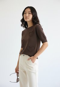 Brązowy krótki sweter z dzianiny z krótkim rękawem, ozdobiony trzema fakturowanymi wzorami serc. Połączony z kremowymi spodniami, trzymany jedną ręką.