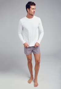 Vit långärmad skjorta i kombination med shorts i rutigt mönster i lila, blått och vitt. Modellen står barfota mot en grå bakgrund.