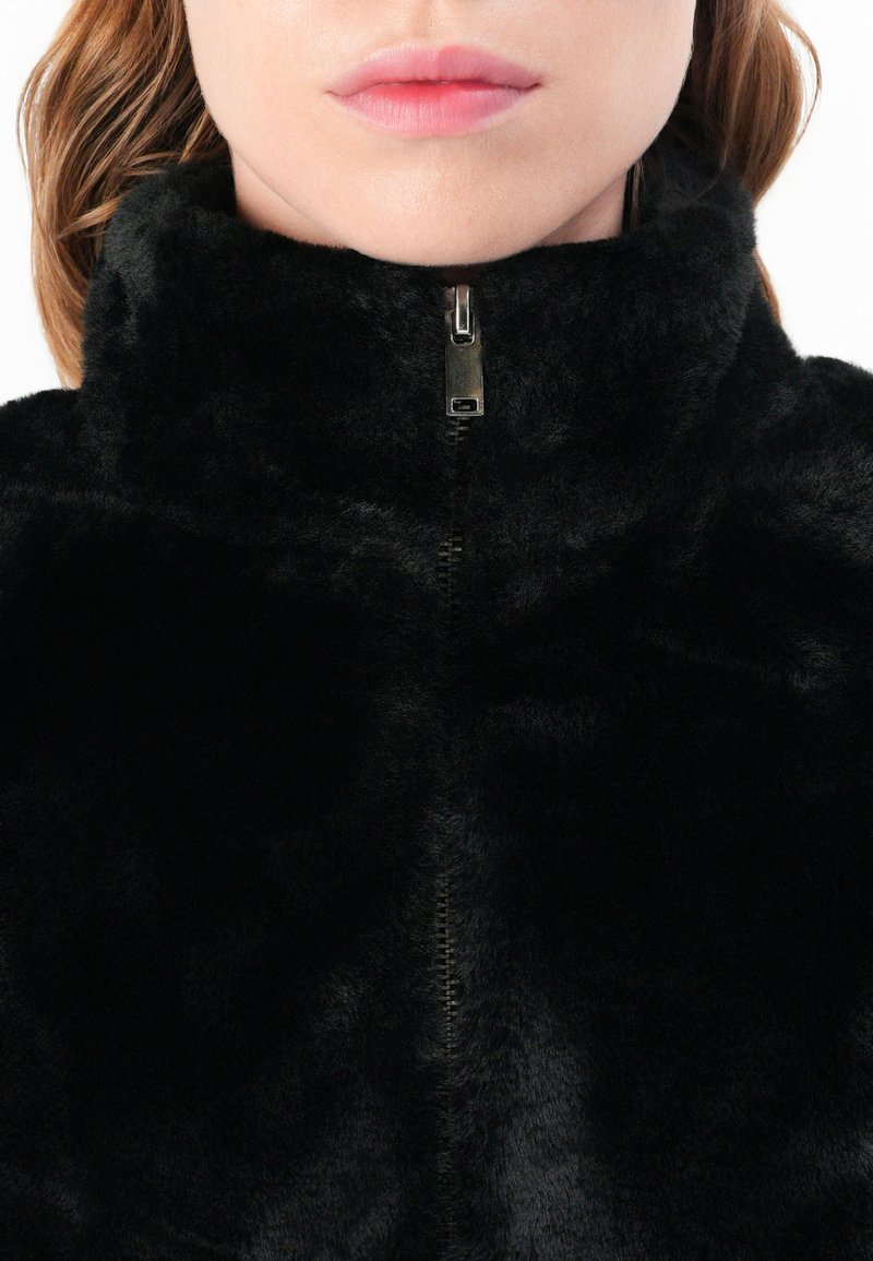 Veste en fausse fourrure noire avec un col montant et une fermeture éclair intégrale. Texture douce et finition lisse, présentant une couleur uniforme et sans motifs.