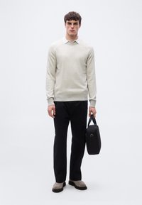 Jeune homme portant un pull beige sur une chemise blanche à col, pantalon noir, bottes grises, tenant une mallette noire devant un fond uni.