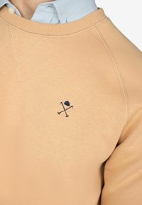 Perzikkleurig sweatshirt met geribbelde ronde hals en geborduurd marineblauw grafisch ontwerp met een hoed en gekruiste stokken op de borst.