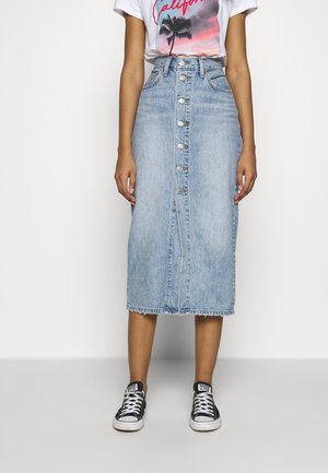 Vrouw draagt een lichtblauwe midi-rok van denim met hoge taille en knopen aan de voorkant, een T-shirt met California-opdruk en zwarte Converse-sneakers.