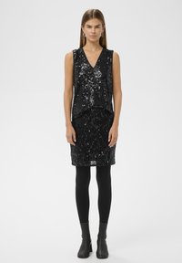 Fekete flitteres ruha V-nyakkivágással, ujjatlan felsőrésszel és testhezálló szoknyával. Fekete harisnyával és bokacsizmával párosítva. Texturált, fényes felület.