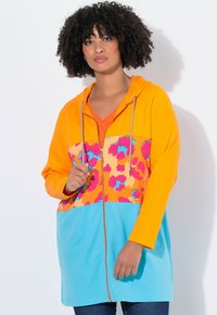 Giacca a felpa colorata con cappuccio arancione, chiusura zip frontale e una sezione inferiore blu con una stampa di leopardo multicolore nella parte superiore.