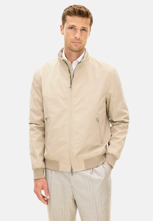 Mand iført beige lynlåstrøje med ribstrikkede manchetter og krave, samt lyse bukser med tynde striber, stående med den ene hånd i lommen.