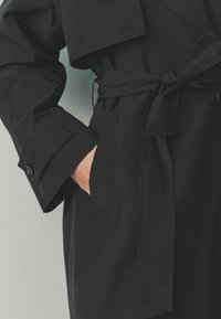 Svart trenchcoat med bälte i midjan, som har bred krage, knäppta ärmslut och sidofickor. Slät textur och skräddarsydd passform.