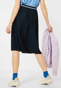 Plisovaná sukně ke kolenům v námořnické barvě, s kontrastním bílým páskem. Model nosí světlé tenisky a modré ponožky, drží lila bundu.