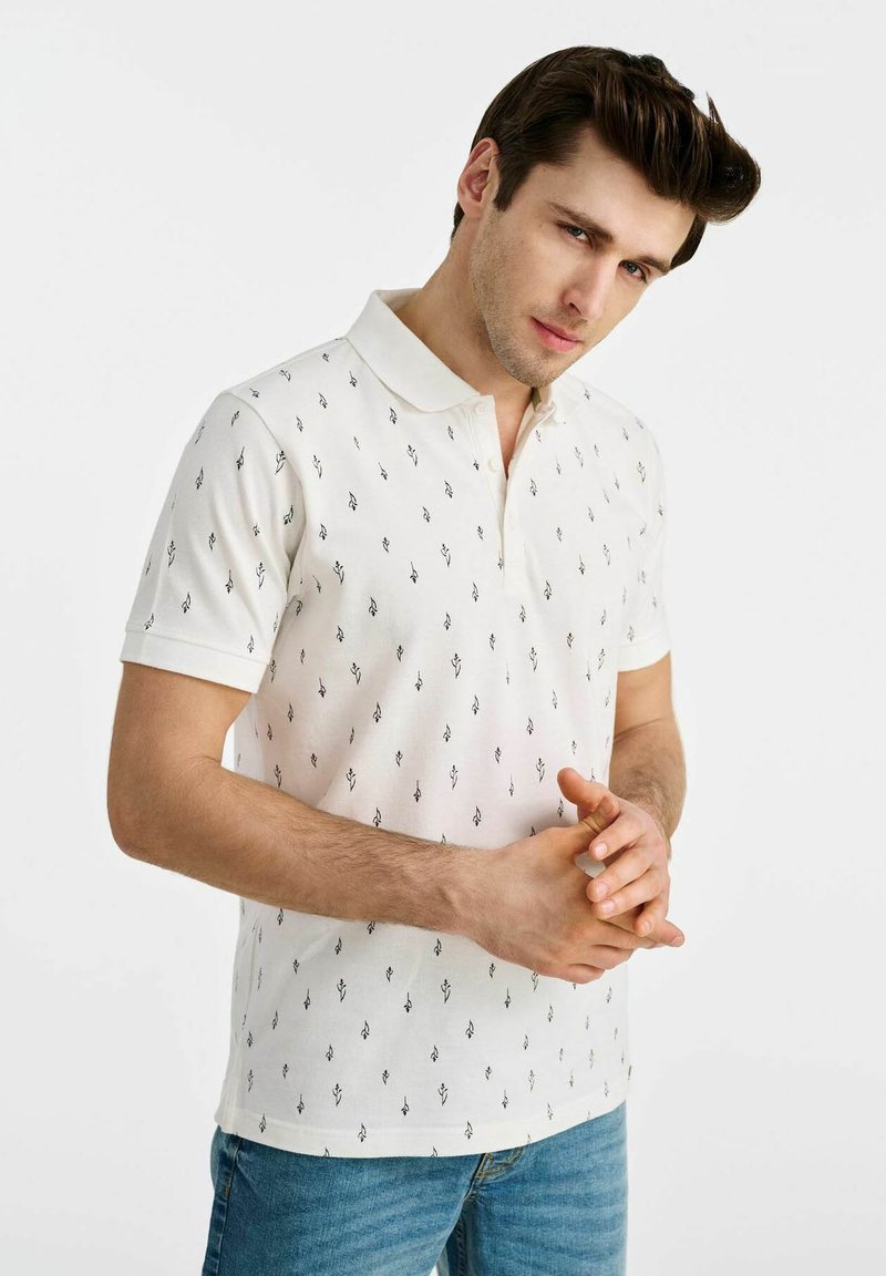 Biała koszulka polo z małymi czarnymi wzorami w strzałki, krótkie rękawy i kontrastowy biały kołnierzyk, w zestawie z niebieskimi dżinsami.