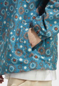 Blå mönstrad jacka med solmotiv i orange och vitt. Har en dold ficka och elastiska manschetter. Lättviktig tyg med lätt textur.