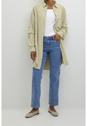 Personne portant une longue chemise boutonnée beige clair sur un haut blanc, un jean bleu droit et des ballerines beiges, debout devant un fond uni.