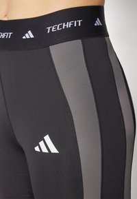 Svarte leggings med Techfit-linning, grå sidepaneler og hvit Adidas-logo. Laget av stretchy, glatt materiale.