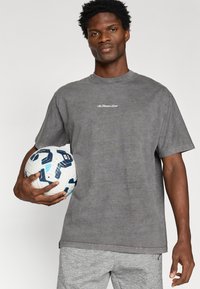 Γκρι βαμβακερό μπλουζάκι με κοντό μανίκι, με λευκό κείμενο στο στήθος. Το μοντέλο κρατά μια λευκή και μπλε κανονική μπάλα ποδοσφαίρου.