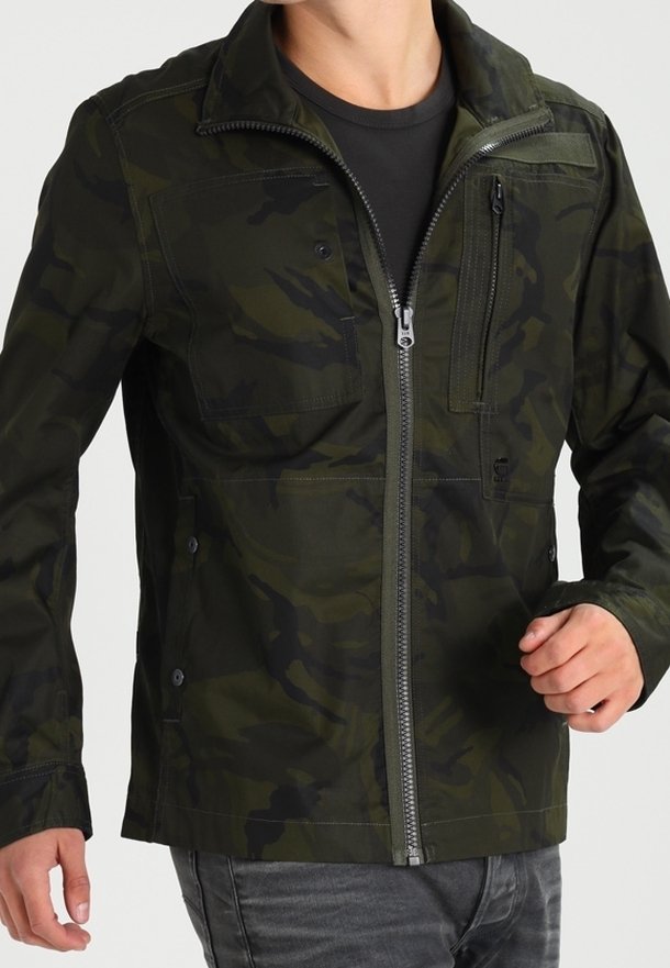 Man som bär en mörkgrön och svart kamouflage zip-up jacka med flera framfickor och en svart tröja under.