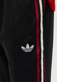 Μαύροι παντελόνες με πλαϊνές ρίγες σε κόκκινο, μαύρο και λευκό, με λευκό λογότυπο Adidas trefoil κοντά στην τσέπη.