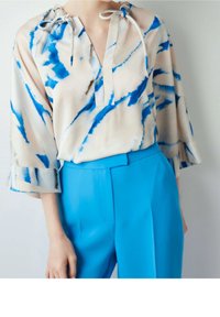 Blusa di seta con motivo astratto blu e beige, caratterizzata da scollo a V, lacci regolabili al collo e maniche a tre quarti voluminose. Abbinata a pantaloni blu brillante.