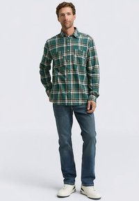 Groen geruit flanellen shirt met witte lijnen, voorzien van een kraag en borstzakken, gecombineerd met een blauwe spijkerbroek en witte sneakers.