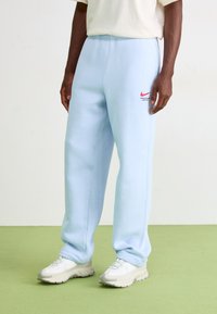 Gaiši zili sporta bikses, kas izgatavotas no mīksta materiāla, ar elastīgu jostu, sānu kabatām un sarkanu Nike logo kreisajā augšstilbā.