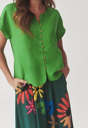 Frau trägt eine hellgrüne kurzärmelige Bluse mit Knöpfen und einen dunklen Rock mit großen bunten Blumenmustern, Hände in den Taschen.
