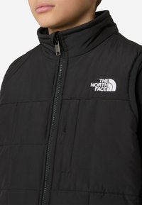 Fekete, bélelt mellény magas gallérral, elöl cipzárral, oldalzsebekkel és hímzett fehér logóval a bal mellrészen. Simább tapintású anyag.