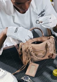 Personne portant des gants blancs inspecte un sac à main beige à l’aide d’une loupe de bijoutier et d’un outil métallique sur une surface de grille noire.