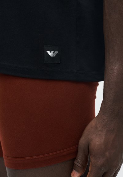 Gros plan d'une personne portant un t-shirt noir avec un patch logo blanc et un short couleur rouille, montrant le bas du torse et la main droite.