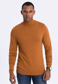 Ockerfarbener Rollkragenpullover aus einem weichen Strickstoff mit langen Ärmeln. Hat eine figurbetonte Silhouette und gerippte Bündchen und Saum. Kombiniert mit blauen Jeans.
