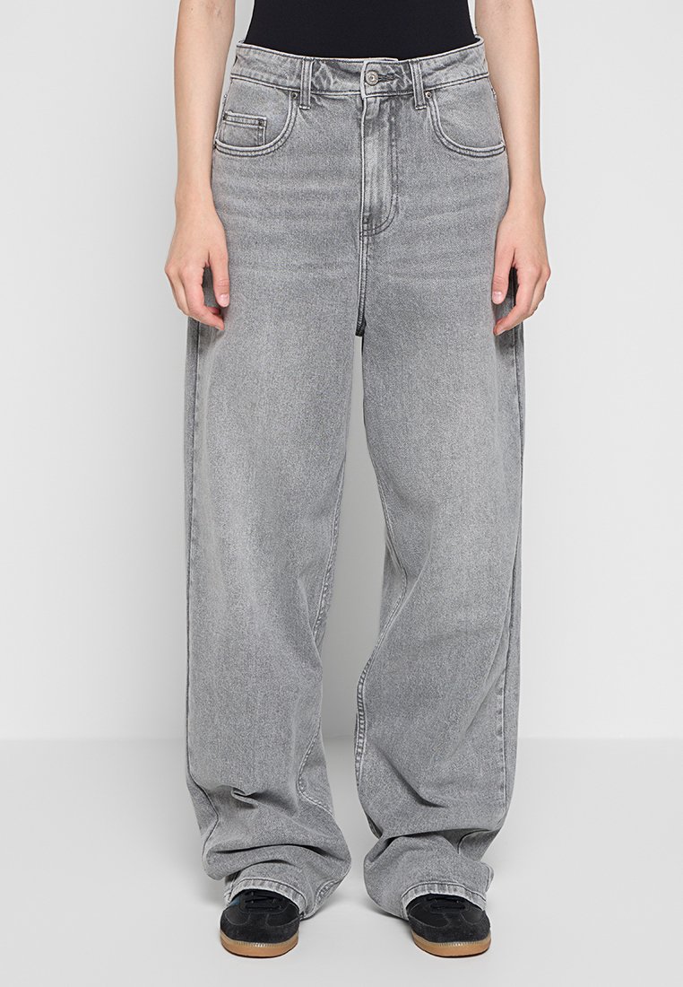 Jeans grigi ampi in denim, con vita alta, grandi tasche frontali e una finitura leggermente sbiadita. Scarpe da ginnastica nere visibili in basso.