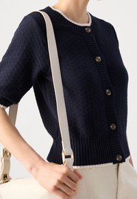 Navy knitted cardigan with textured design, short sleeves, and ribbed hem. Features light-colored trim and brown buttons.