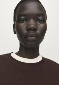 Hanorac maro închis cu guler alb cu dungi, purtat de o persoană cu piele închisă la culoare, pe un fundal neutru. Prim-plan cu fața.