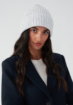 Light gray knitted beanie with ribbed texture, featuring a rounded top and foldover cuff, paired with a dark navy coat and white sweater.
