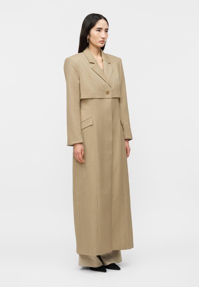 Γυναίκα με ίσια σκούρα μαλλιά που φοράει μακρύ, μπεζ, ραμμένο παλτό και αντίστοιχο παντελόνι με φαρδιά μπατζάκια, στέκεται μπροστά σε ένα απλό φωτεινό φόντο.