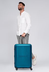 Hartschalenkoffer in Teal mit vertikalen Rillen, ausziehbarem Griff und vier Rollen. Glatte Textur und zentriertes Logo auf der Vorderseite.