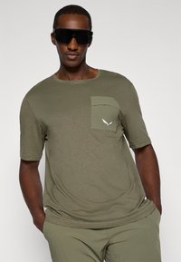 Ελαιοπράσινο μπλουζάκι με στρογγυλή λαιμόκοψη, κοντά μανίκια και υφασμάτινη τσέπη. Διαθέτει λευκό λογότυπο πάνω από την τσέπη.