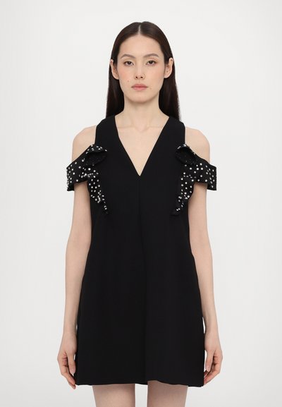 Zwarte jurk met een V-hals en een koud-schouder ontwerp, voorzien van ruffles met witte stippen. Soepele textuur, korte lengte.