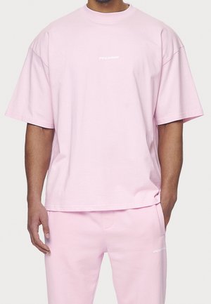 Mand iført lyserød oversized t-shirt og matchende bukser med lille hvid "PEGADOR" tekst på bryst og lår.