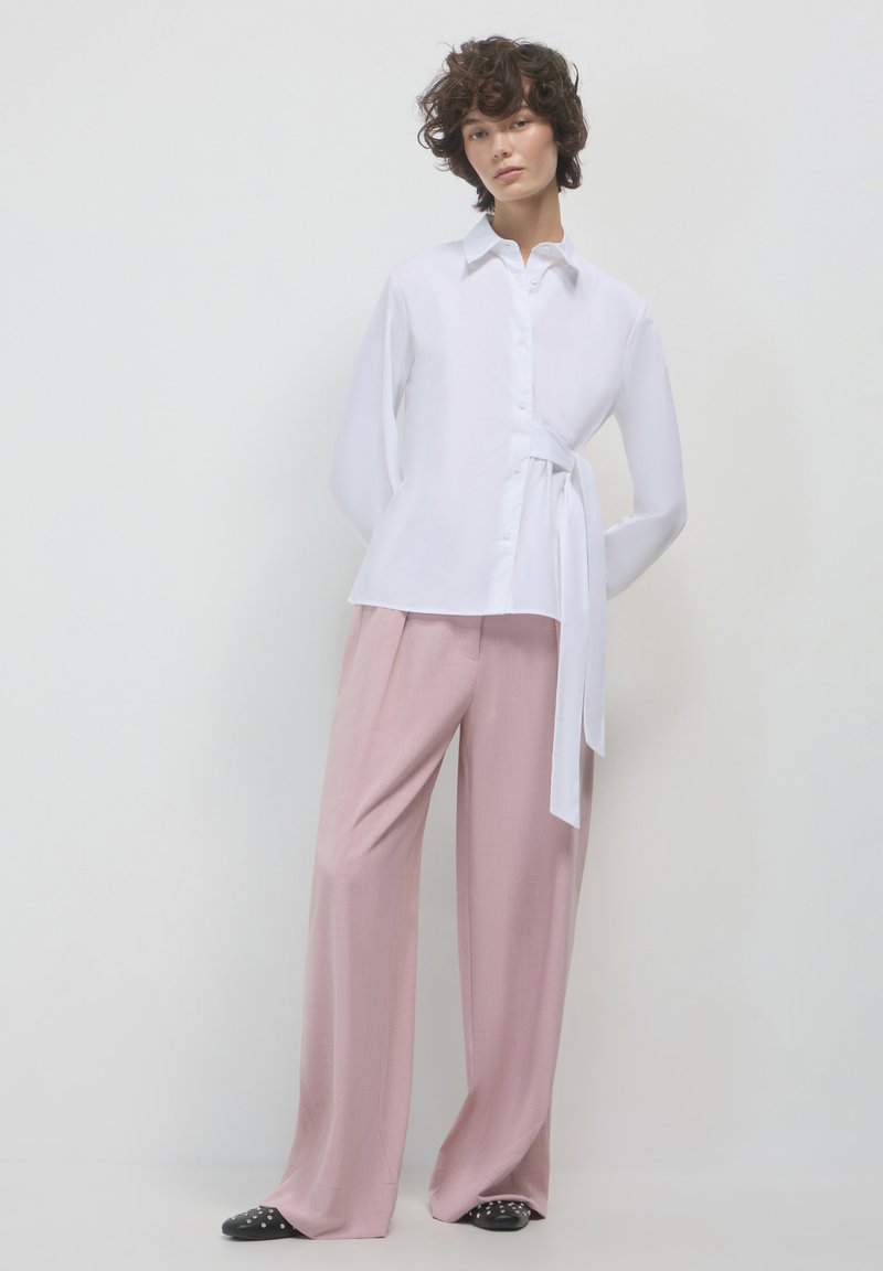 Persona che indossa una camicia bianca con bottoni, cravatta laterale e pantaloni larghi di un rosa chiaro, in piedi contro uno sfondo chiaro e uniforme.