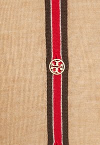 Beige Strickstoff mit einem roten und braunen Streifen. Ein goldener runder Logo-Knopf verleiht dem Zentrum des Streifens das gewisse Etwas.