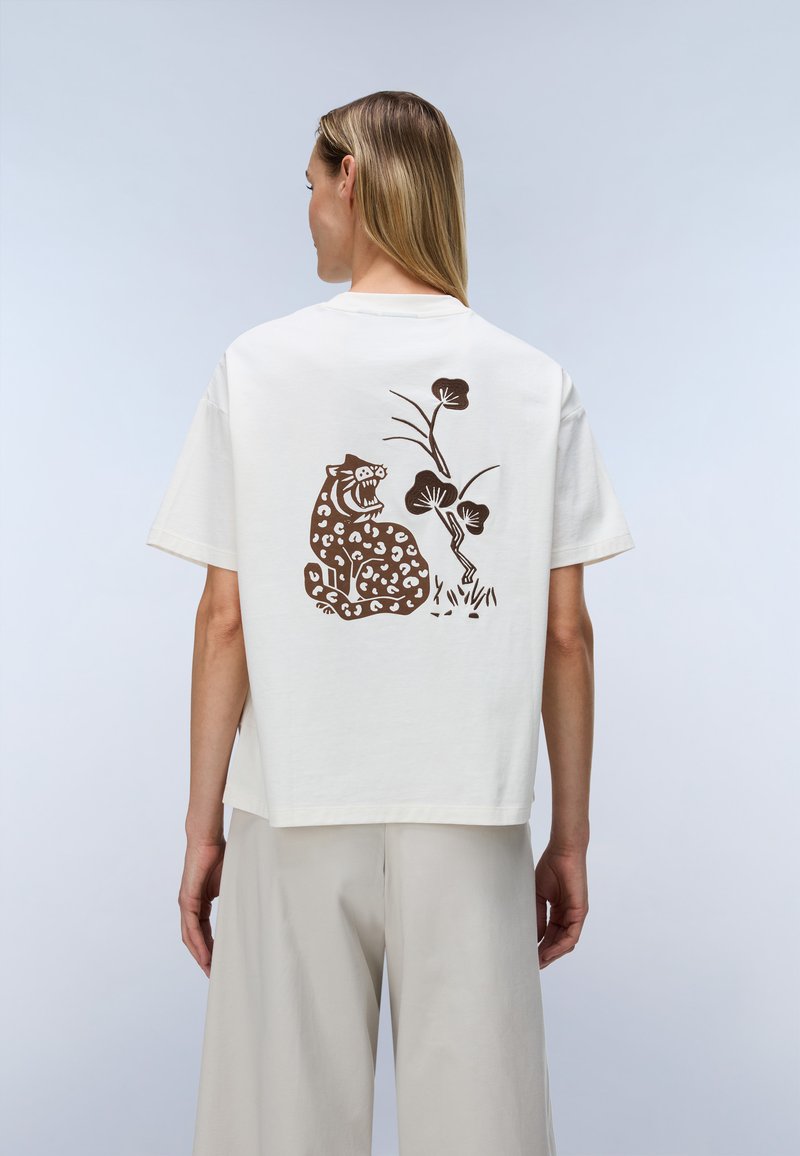 Γυναίκα που φοράει ένα λευκό t-shirt με καφέ γραφικό ενός βρυχώμενου λεοπάρδαλου και φυτών στην πλάτη, σε συνδυασμό με ανοιχτό μπεζ φαρδιά παντελόνια.