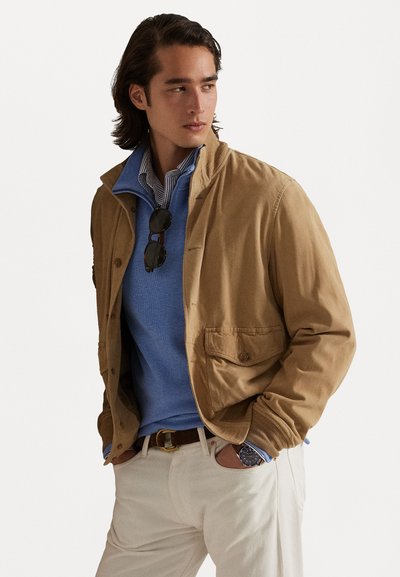 Casaco de algodão bege com colarinho com fecho, bolsos frontais e botões castanhos. Usado sobre um suéter azul claro e calças brancas.