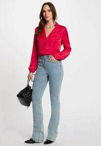 Blusa di raso rosso brillante con due tasche frontali, abbinata a jeans a palazzo blu chiaro a vita alta. Tiene in mano una piccola borsa nera.