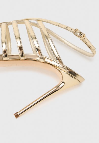 Χρυσό μεταλλικό πέδιλο με ψηλό τακούνι που διαθέτει κάθετες λουριά, λεπτό λουρί στον αστράγαλο με αγκράφα και μυτερό σχέδιο στα δάχτυλα. Λεία υφή.