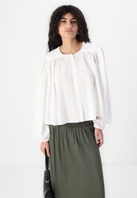 Blouse blanche à manches longues avec un devant à boutons et des détails froncés, associée à une jupe à taille élastique de couleur vert foncé. Le modèle tient un sac noir.